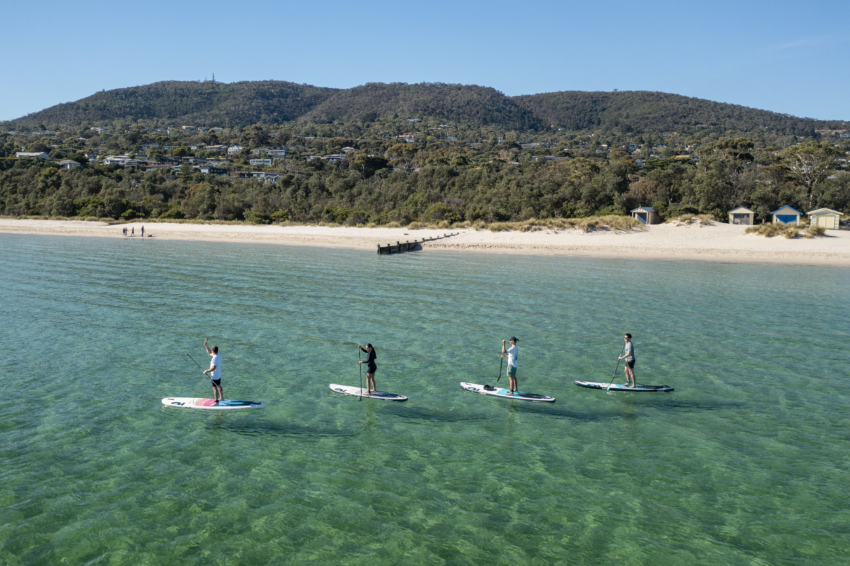 Stand Up Paddleboarding instructor paddling with small group in turquoise blue waters with beach and bathing boxes in the background.