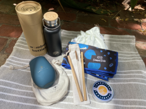 Image of water bottle, reusable coffee cup, bamboo straws, reef-safe sunscreen in a tin, and a towel